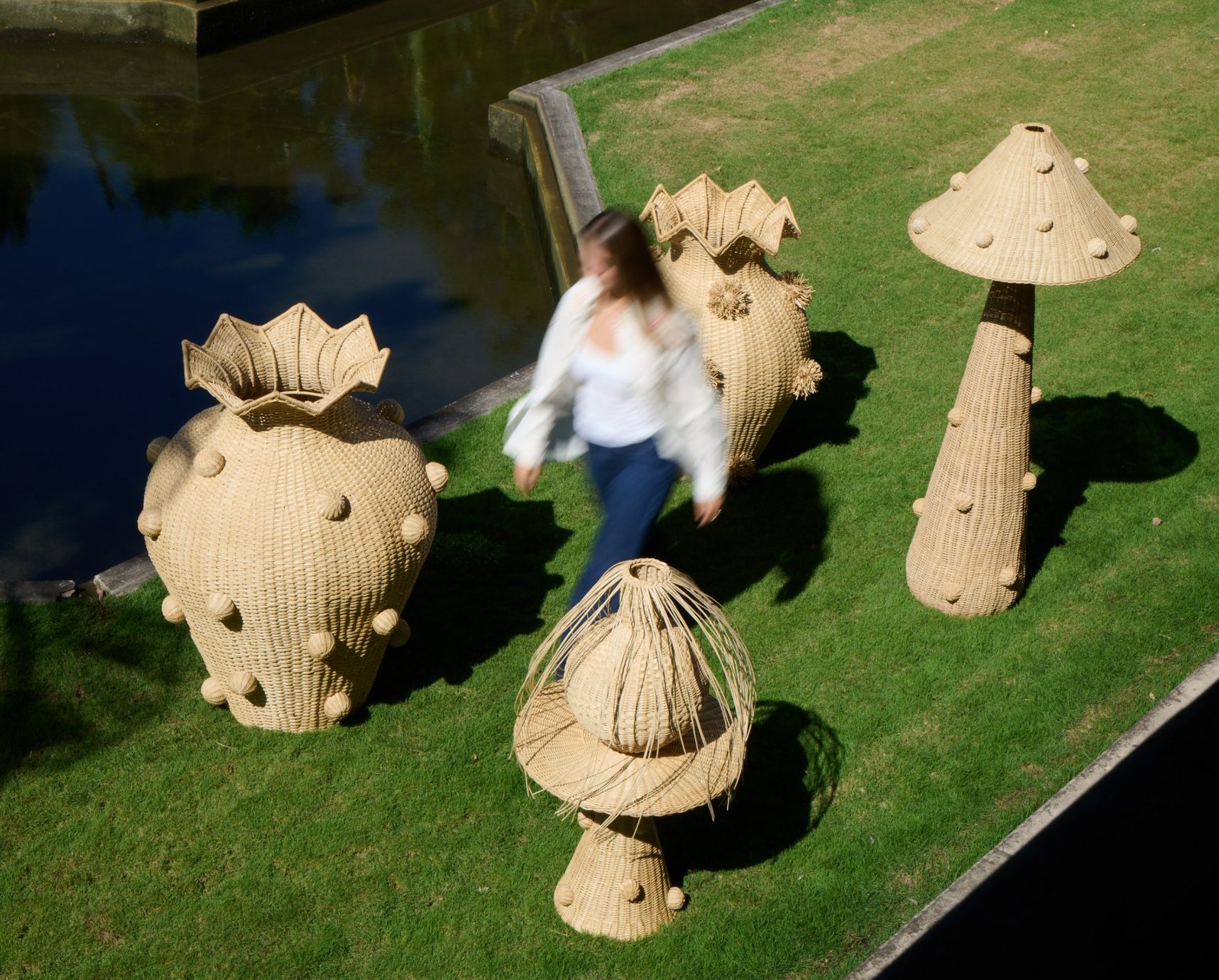Large handwoven Cálamo vases and mushroom lamps on a lawn beside a reflecting pool
